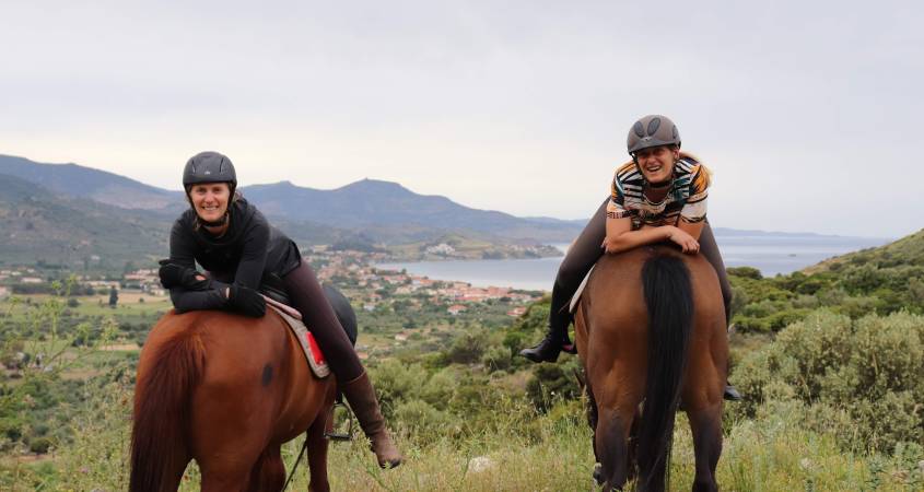 Two adventurous women have fun during a break of riding horses in Greece. While the horses face towards the town of Petra on Lesvos, the women sit backwards in the saddle resting their arms on the horses hips.